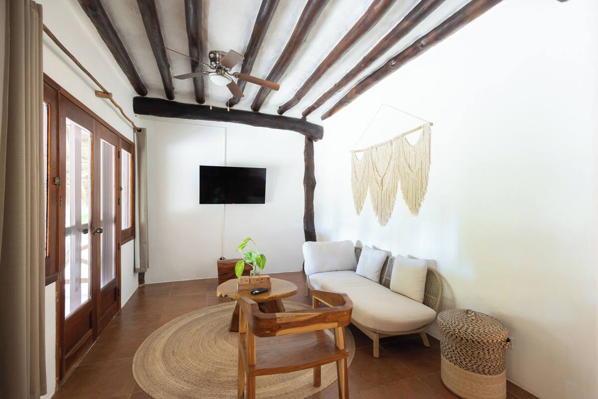 A living room with a wooden ceiling, a flat-screen TV mounted on the wall, a sofa, a coffee table, and a wooden chair.