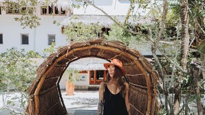 A person walking through a rustic archway with a thatched roof, surrounded by greenery and a building with a red roof in the background. - Las Nubes de Holbox (Isla Holbox)