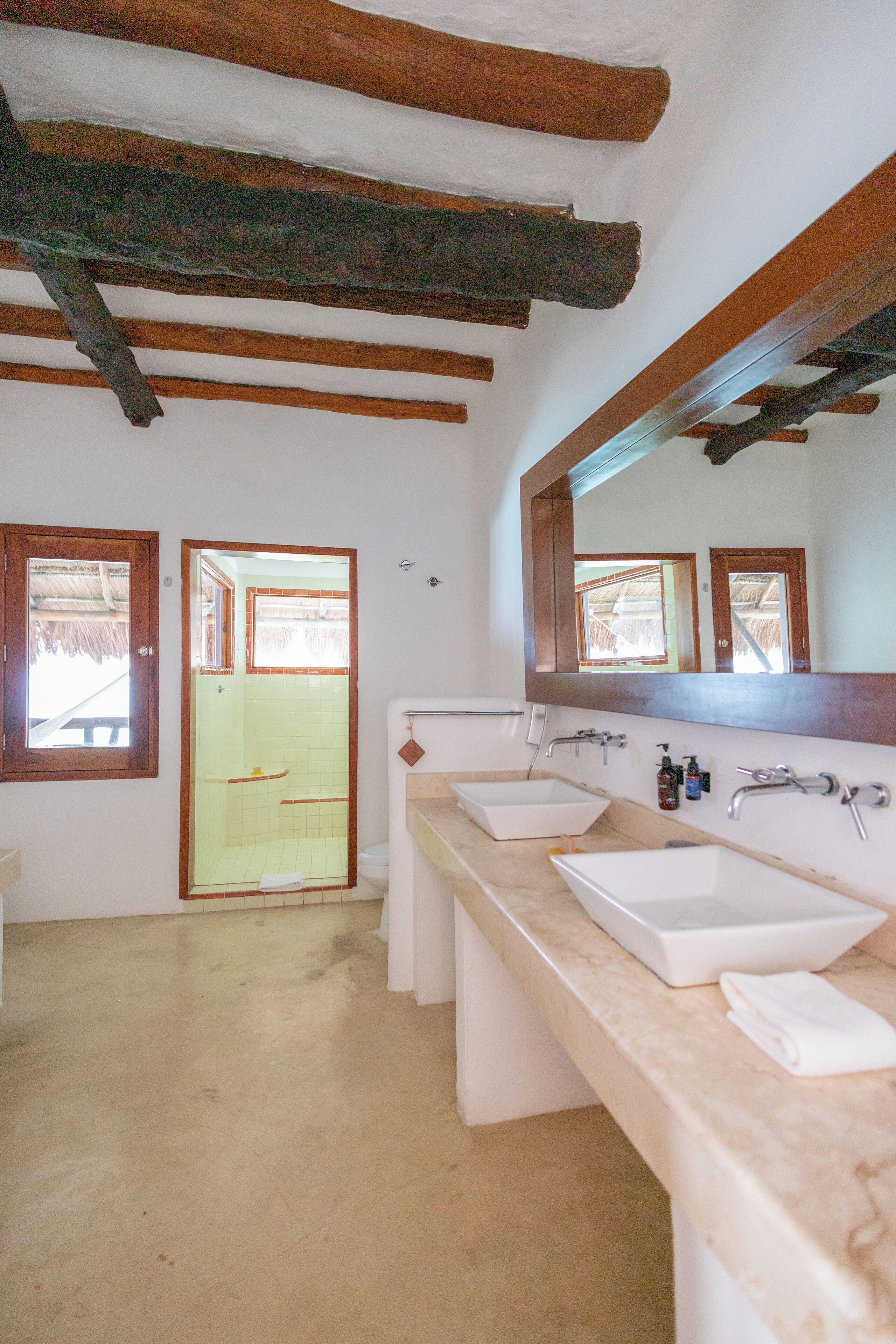 Un baño con dos lavamanos, un espejo grande, vigas de madera en el techo y una puerta que da al exterior.