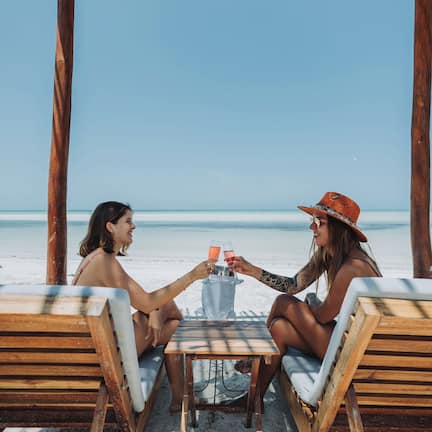 Two people seated at a wooden table with a drink, under a wooden pergola on a beach.