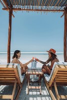 Duas pessoas sentadas à mesa de madeira com uma bebida, sob uma pérgula de madeira na praia.