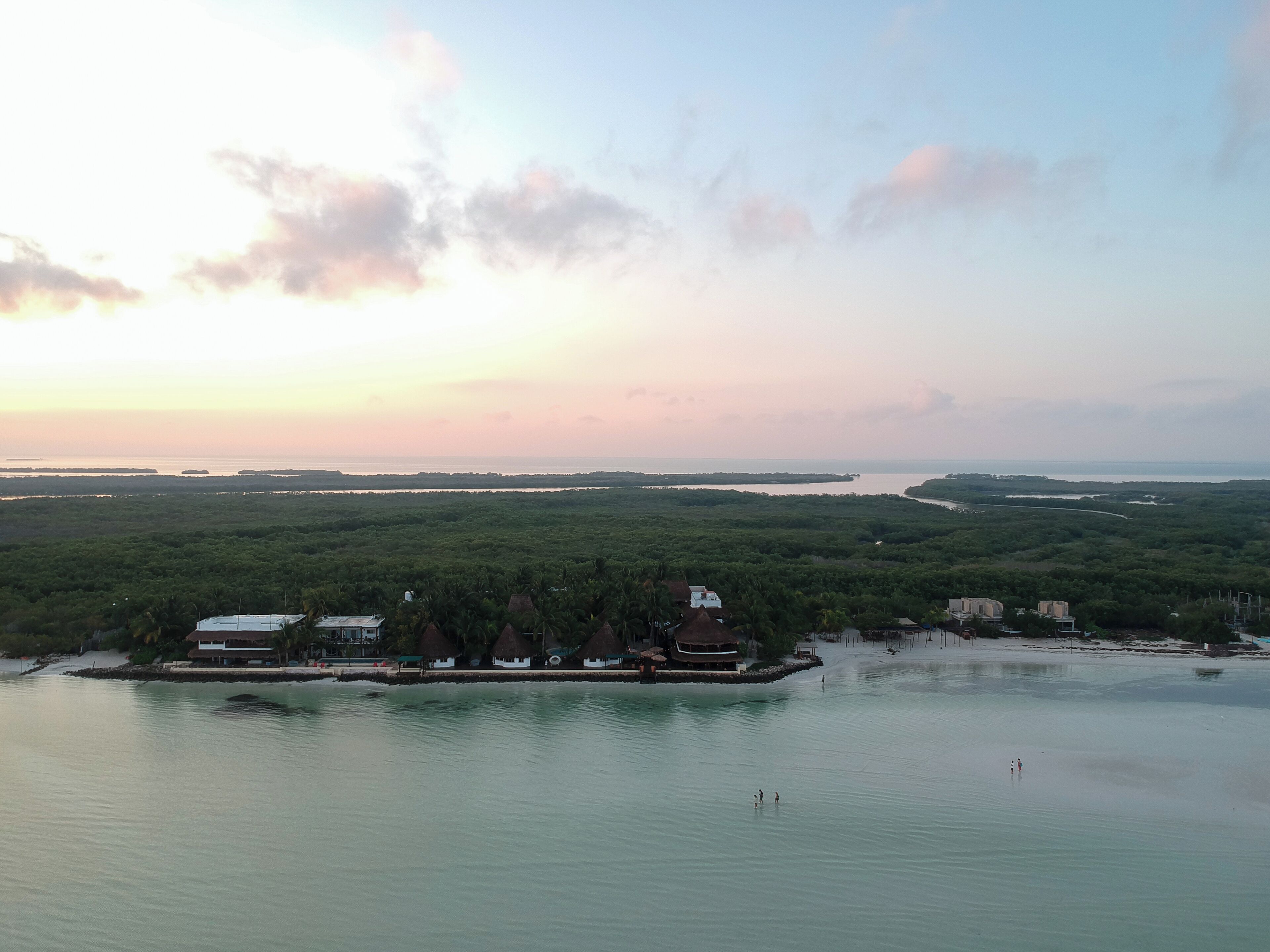 A coastal landscape with a water body, dense greenery, and a few buildings with thatched roofs.