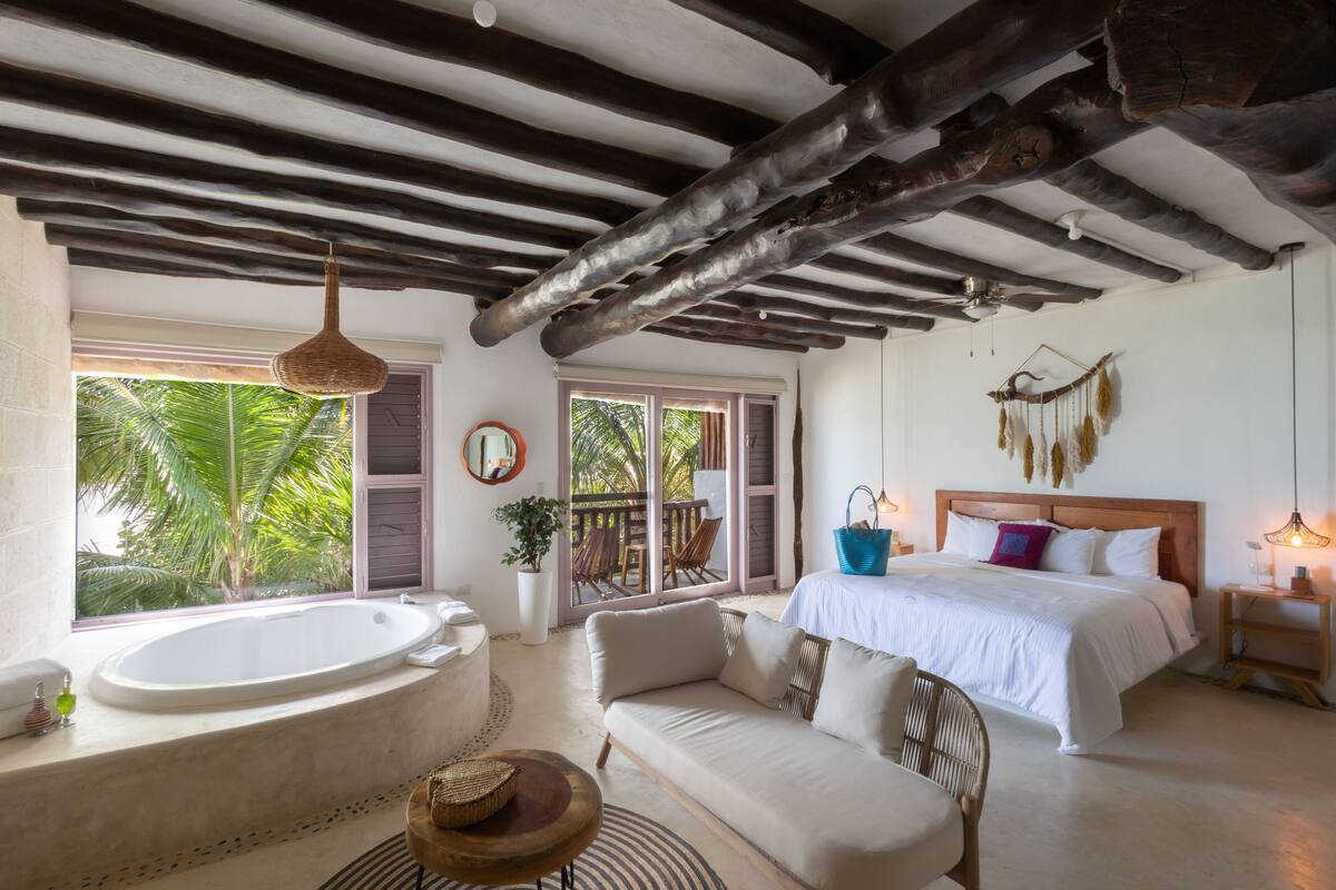 A spacious room with a large bathtub, a comfortable sofa, a bed with white bedding, and a wooden ceiling.