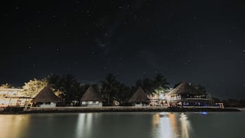 Un hotel frente a la playa con estructuras de techo de paja, una piscina tranquila y un cielo nocturno estrellado.