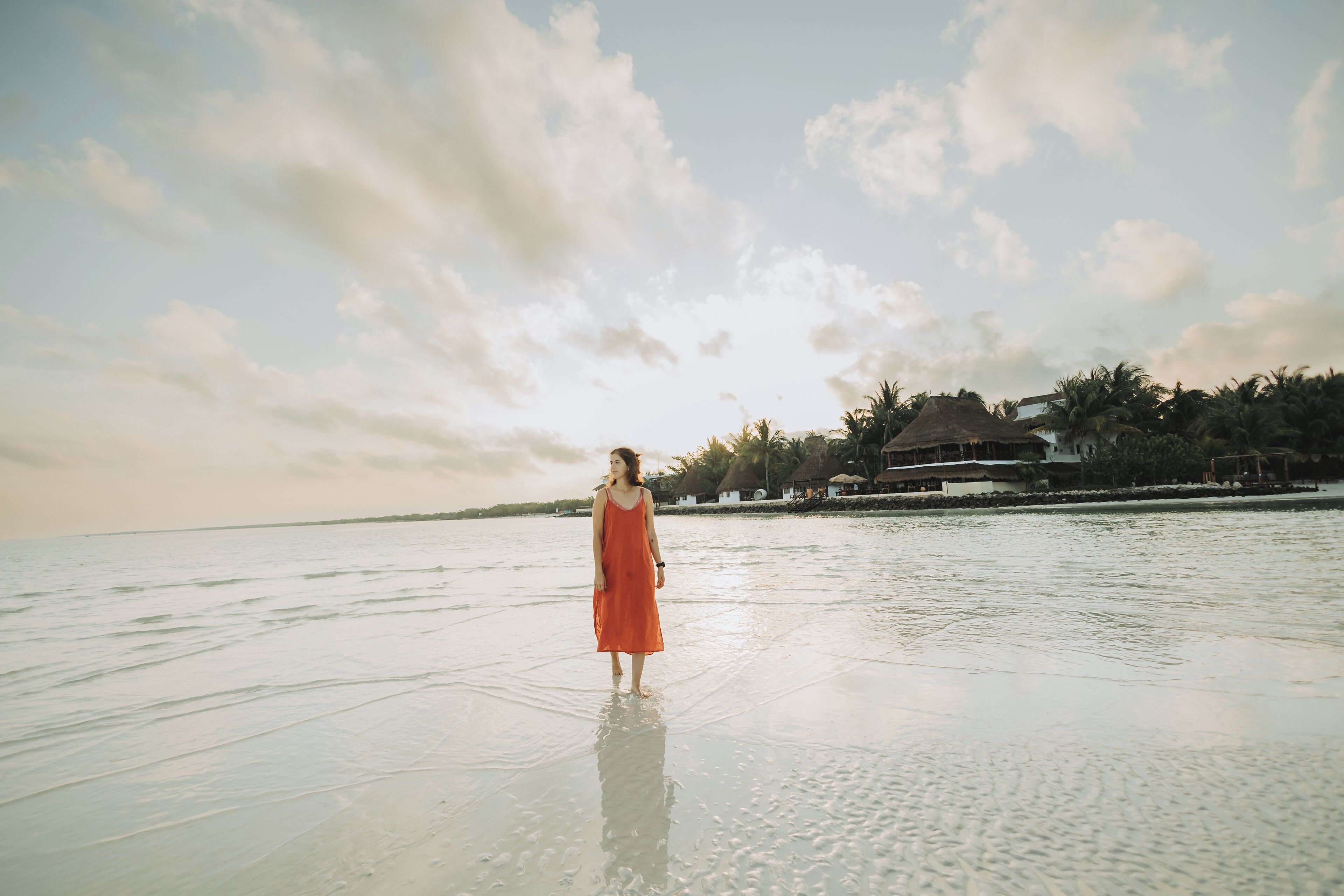 A person in a red dress standing on a beach with a calm sea and a clear sky.