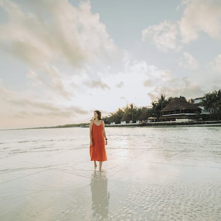 A person in a red dress standing on a beach with a calm sea and a clear sky.