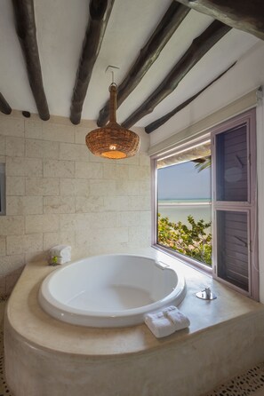 Un baño con una bañera grande, una lámpara de mimbre y una ventana con vista a la playa.