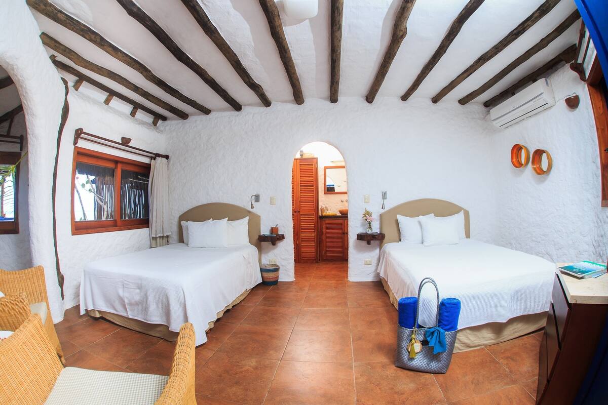 A room with two beds, a wooden ceiling, a window with curtains, a door, and a small table with a handbag.