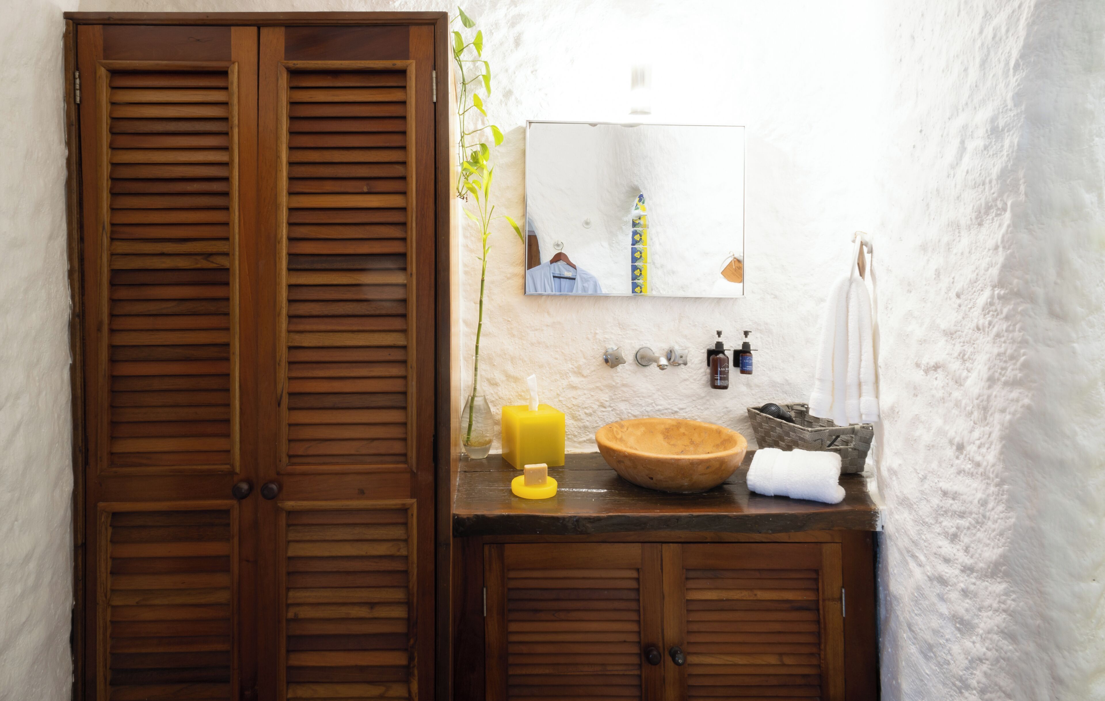 A bathroom with wooden shutters, a mirror, a sink with toiletries, and a towel rack.