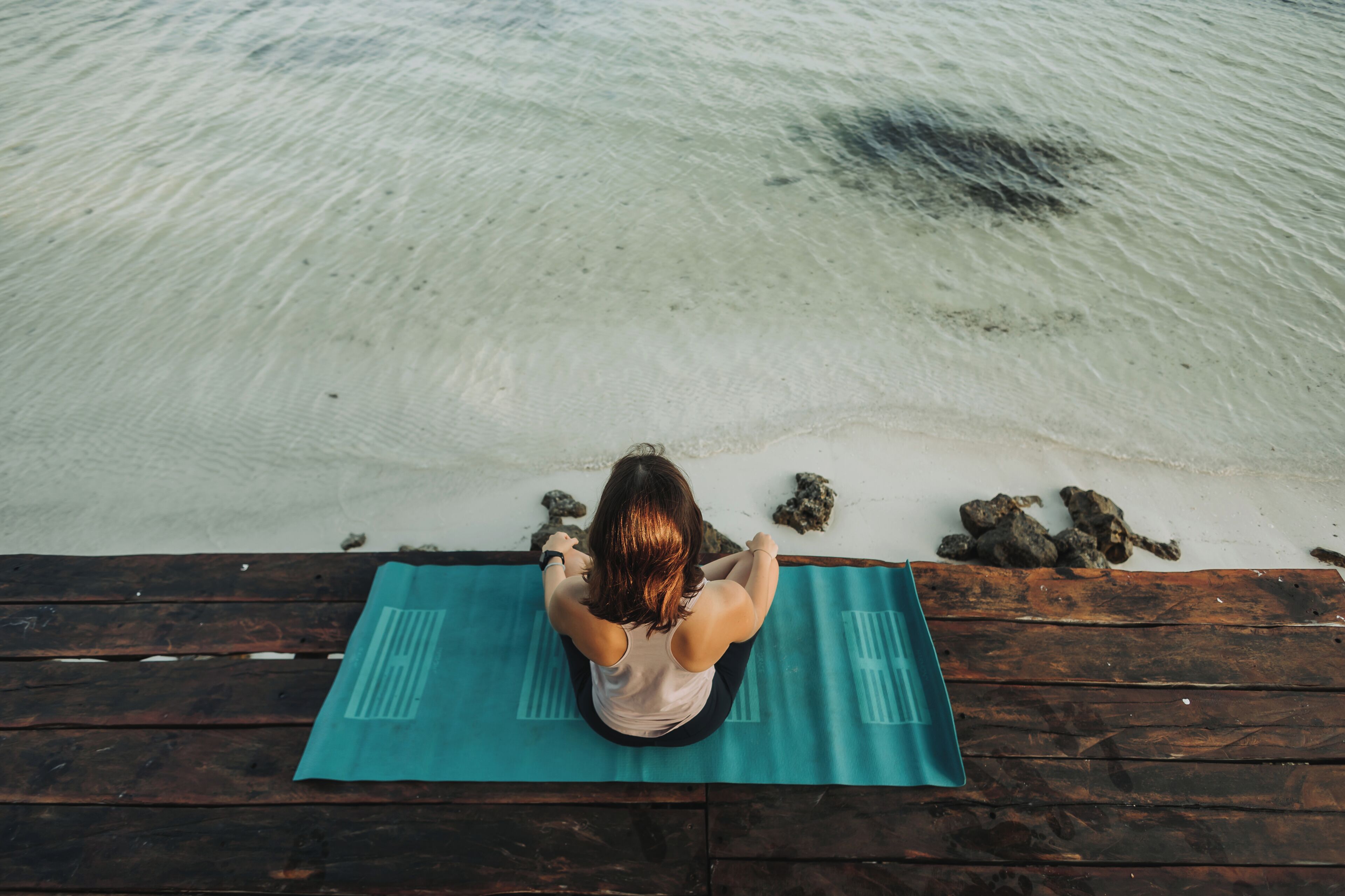 A person sitting on a yoga mat on a wooden dock by a clear beach.
