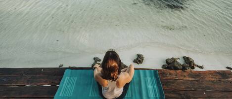 A person sitting on a yoga mat on a wooden dock by a clear beach.