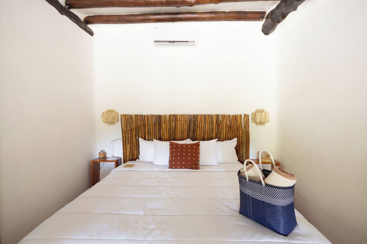 A neatly made bed with white linens, a wooden headboard with a patterned drape, a bedside lamp, and a blue woven bag on the bed.
