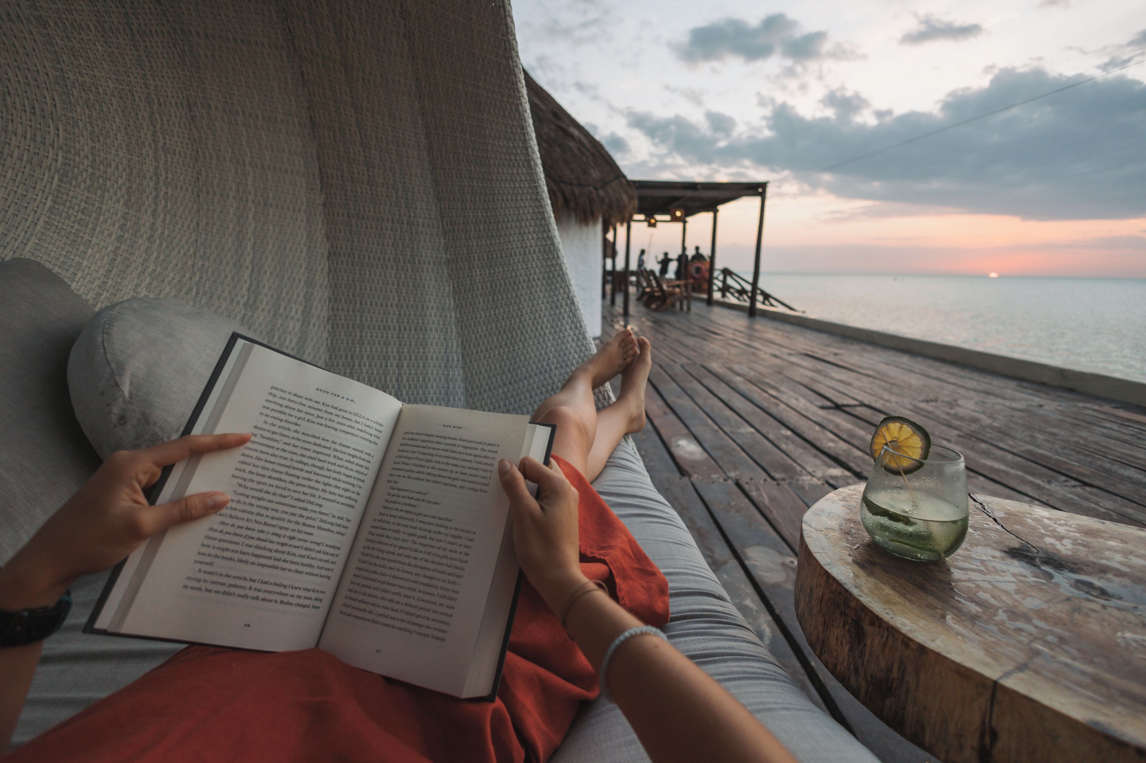 A person reading a book on a wooden deck with a view of the sea and a thatched-roof structure in the background.