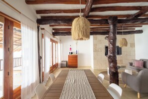 Un comedor con una mesa de madera, sillas blancas, una lámpara colgante tejida y una pared de piedra al fondo.