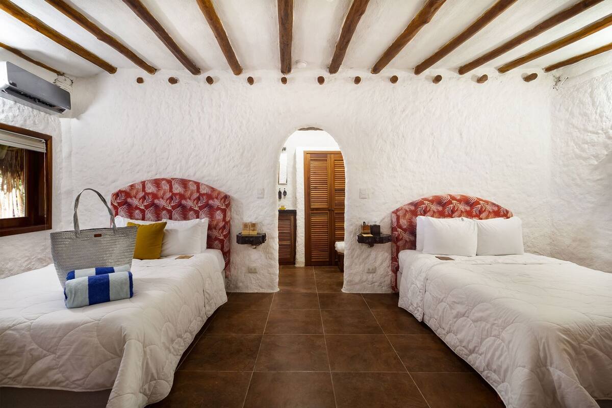 A room with two beds, a large woven bag, and a wooden door in the background.