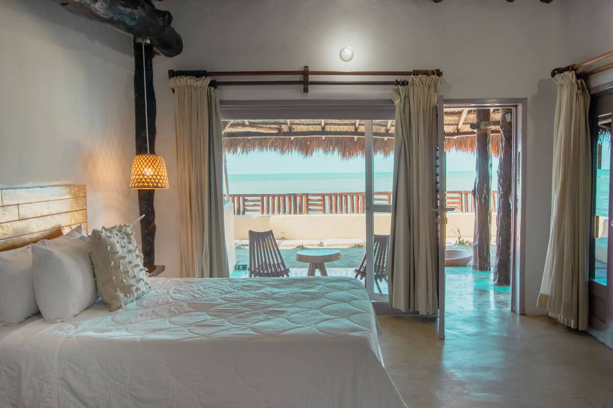 A bedroom with a large bed, a nightstand, a lamp, and a view of the beach through an open door.