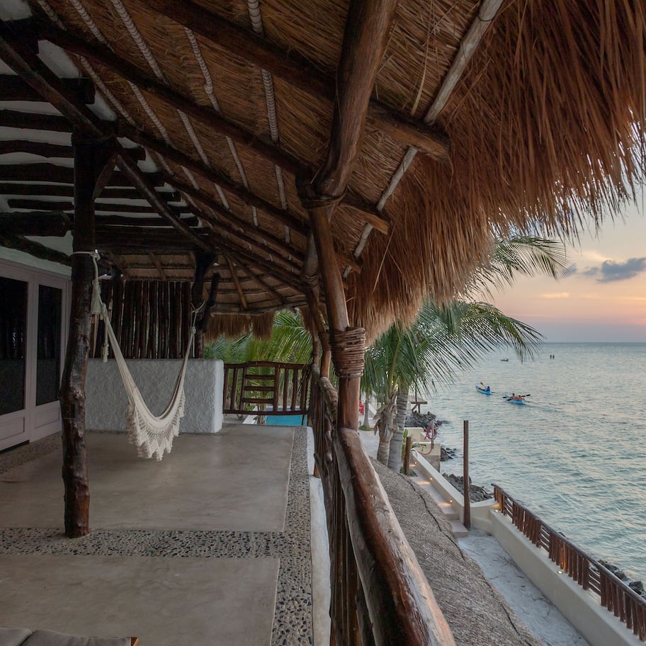 A beachside resort with a thatched roof structure, a hammock, and a view of the sunset over the ocean.