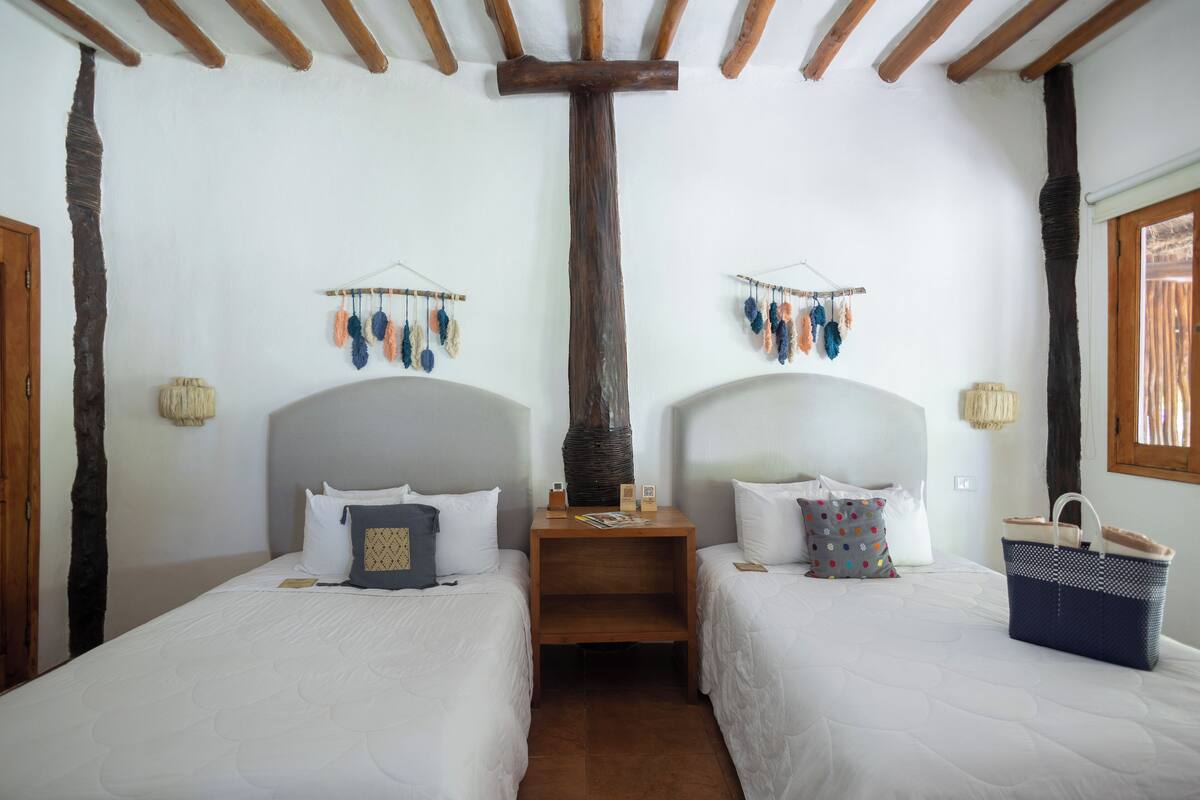 A room with two beds, wooden beams on the ceiling, a wooden nightstand, and a basket on the right side.