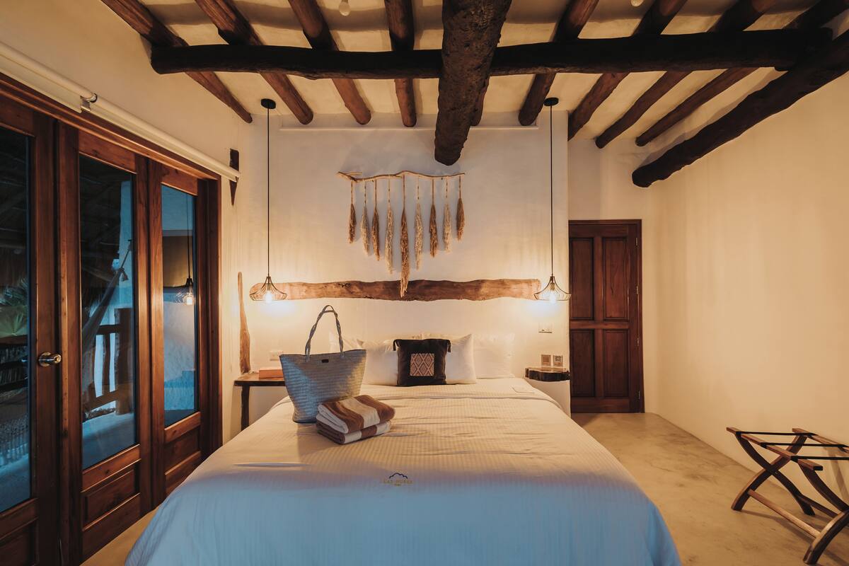 A bedroom with a large bed, wooden ceiling beams, a hanging decoration, and a wooden door.