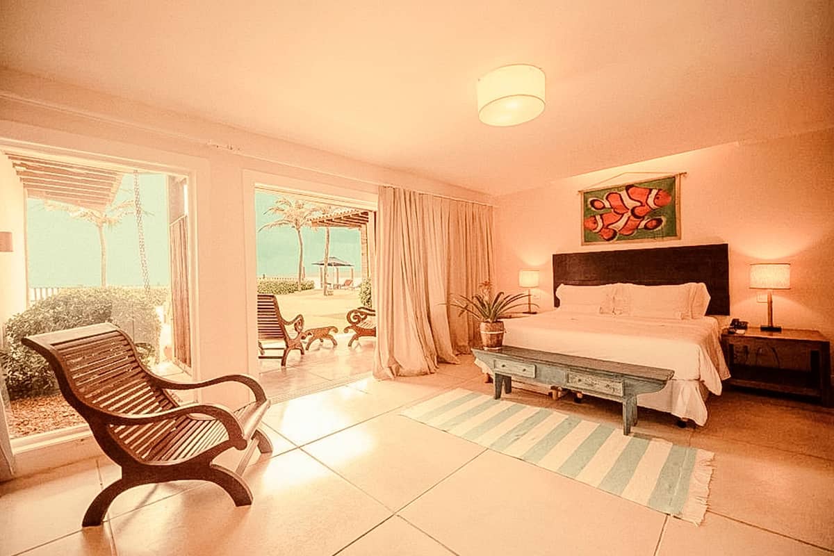 A hotel room with a bed, a bench, a chair, a nightstand, a lamp, and a view of the outdoors.