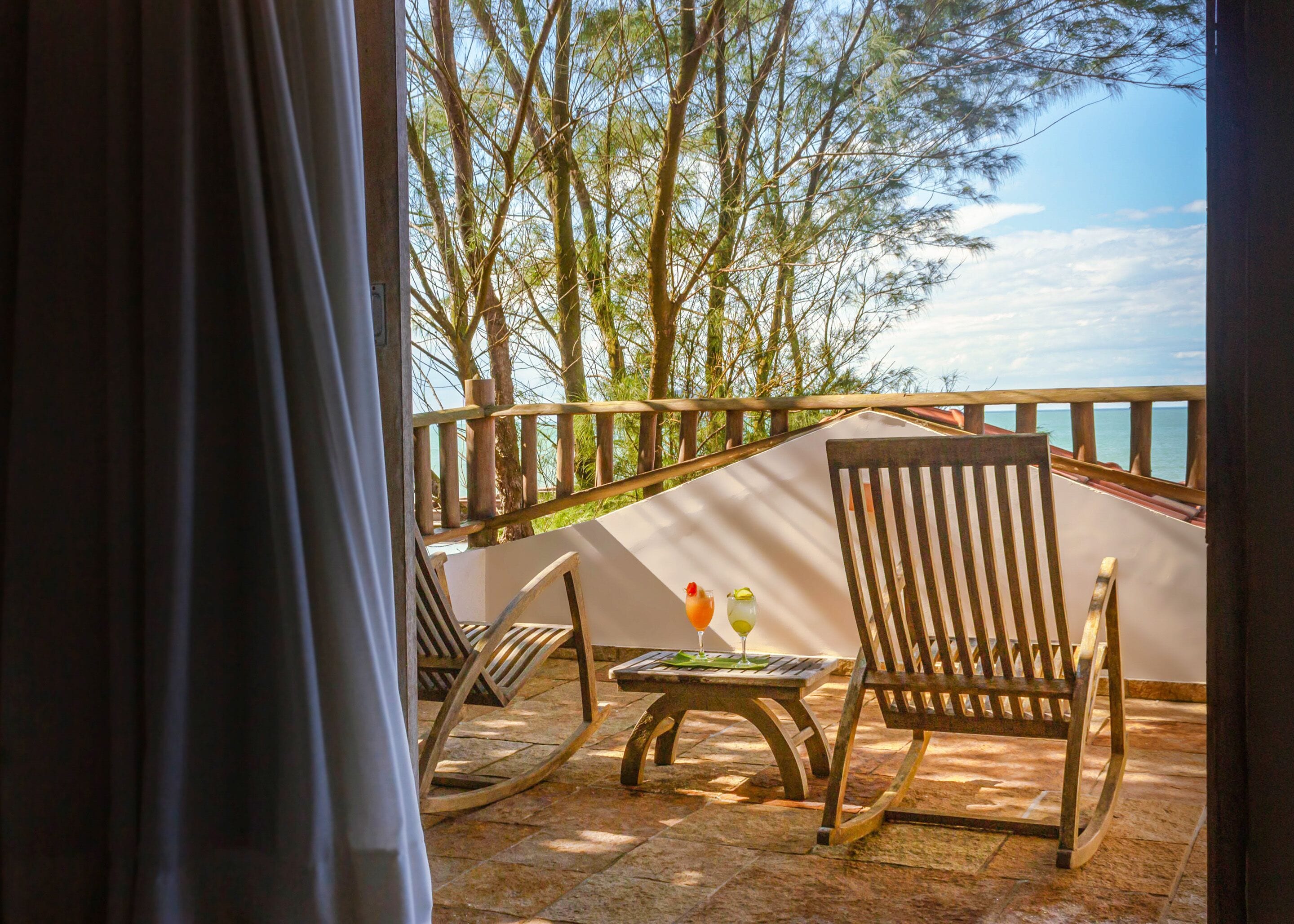 A balcony with wooden chairs and a table, overlooking a body of water and trees.