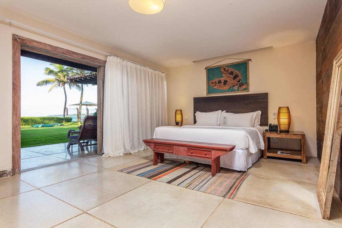 A bedroom with a bed, bedside lamps, a wooden bench, a striped rug, and a view of a lawn and palm trees through a large window.