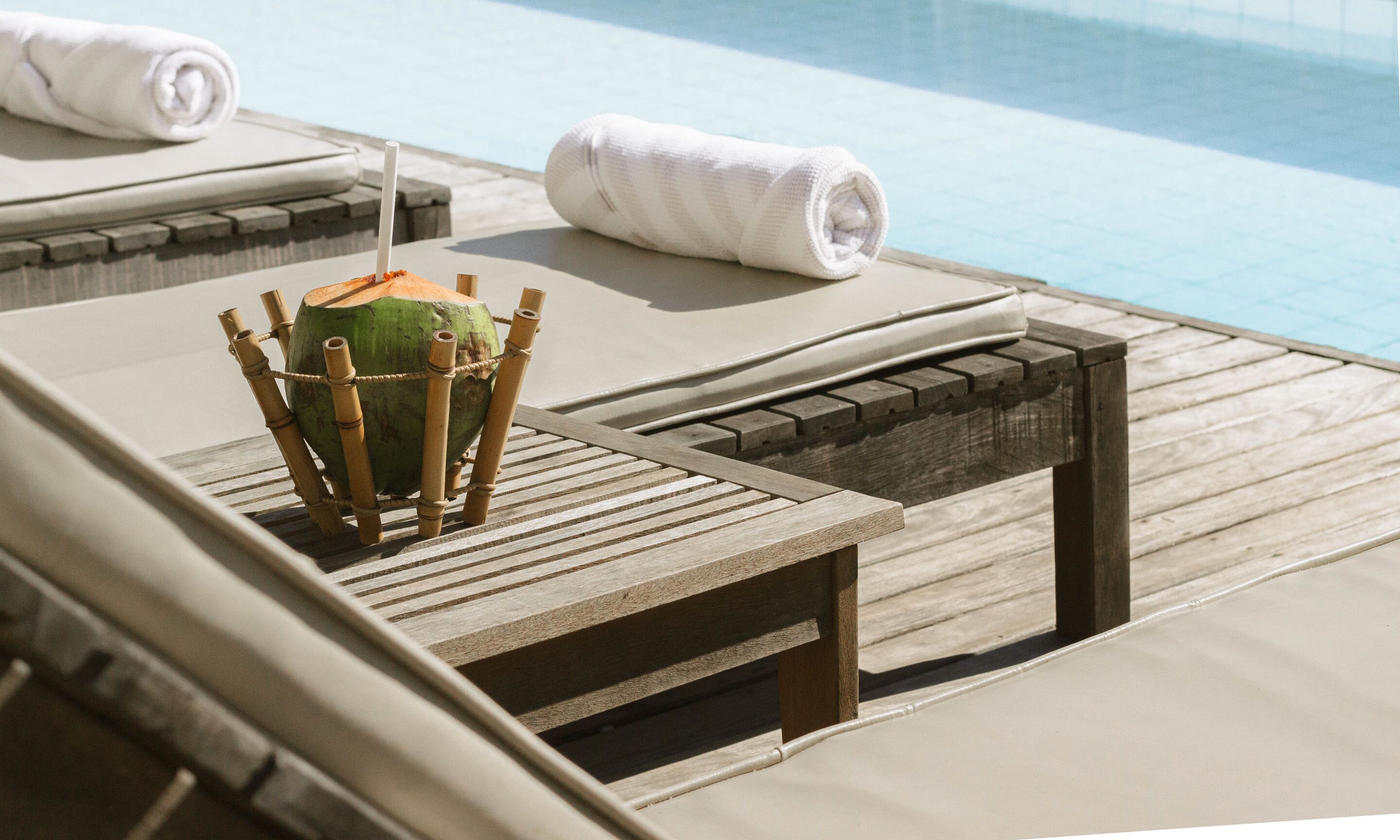 Un área de descanso junto a la piscina con tumbonas de madera, una silla de bambú y una toalla blanca.