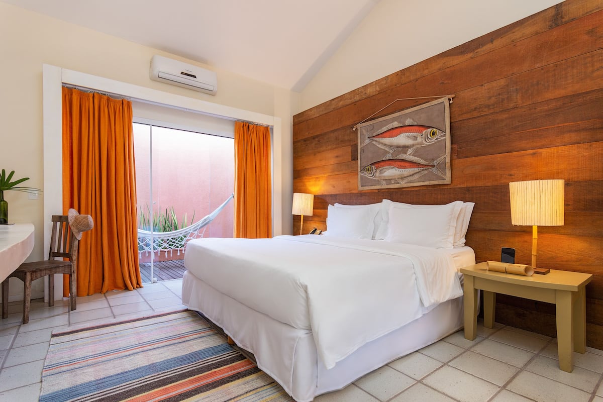 A hotel room with a bed, a nightstand, a chair, a table, and a window with curtains.