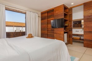Habitación de hotel con una cama, un televisor montado en una pared de madera, una ventana con cortinas y vistas a un edificio con tejado de tejas.