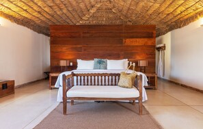A bedroom with a wooden headboard, a bed with white linens, a bench with a woven bag, and a nightstand with a lamp.