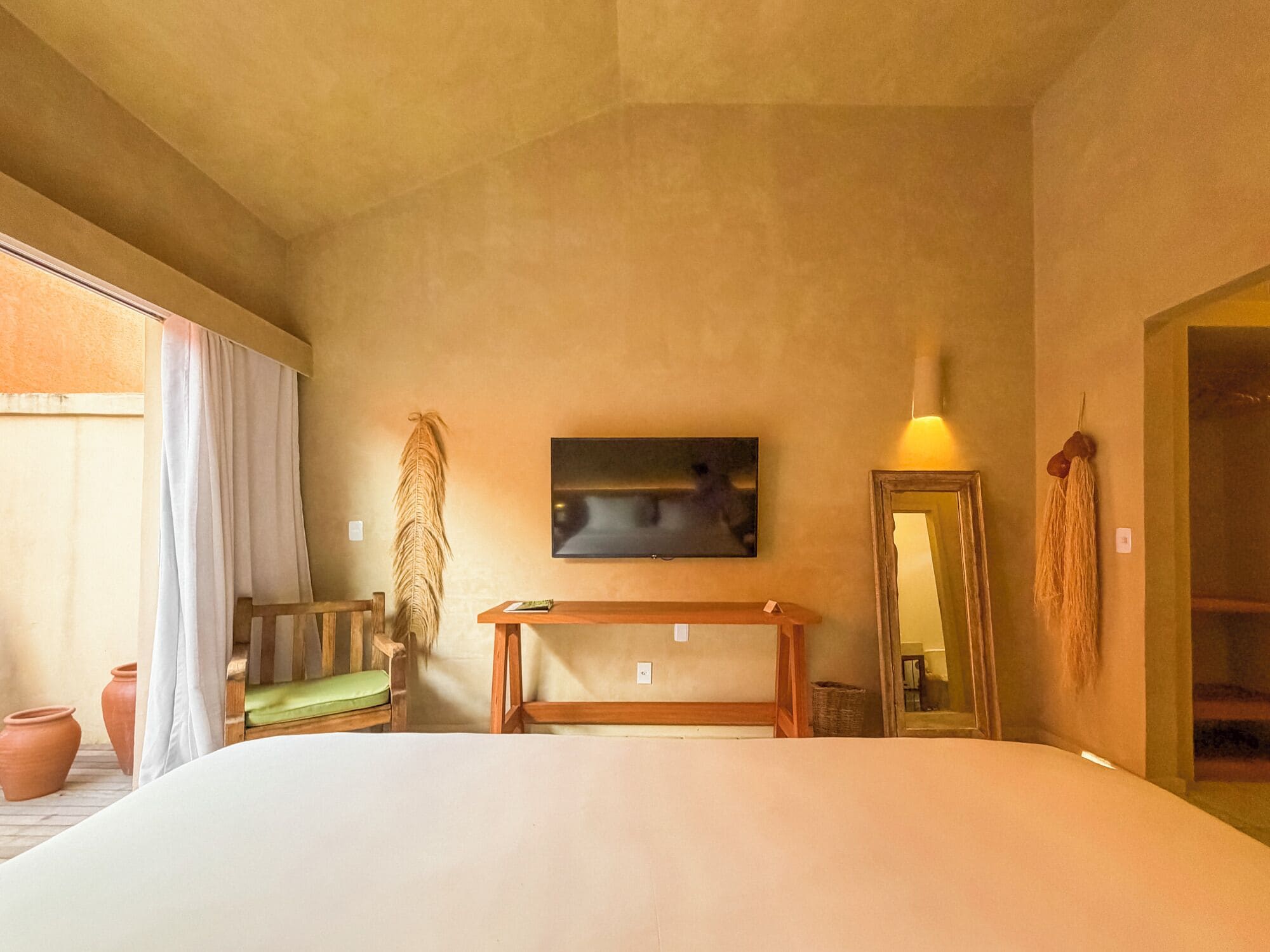 Un dormitorio con una cama, una mesita de noche de madera, un espejo, un televisor montado en la pared y una ventana con cortinas.