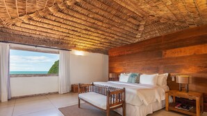 A bedroom with a wooden ceiling, a bed with white bedding, a wooden bench, a nightstand with a lamp, and a view of the sea through a window. - Villa Rasa (Búzios)