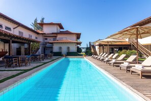 Una piscina con sillas de descanso y una zona cubierta, rodeada de edificios con techos de tejas y una escalera.