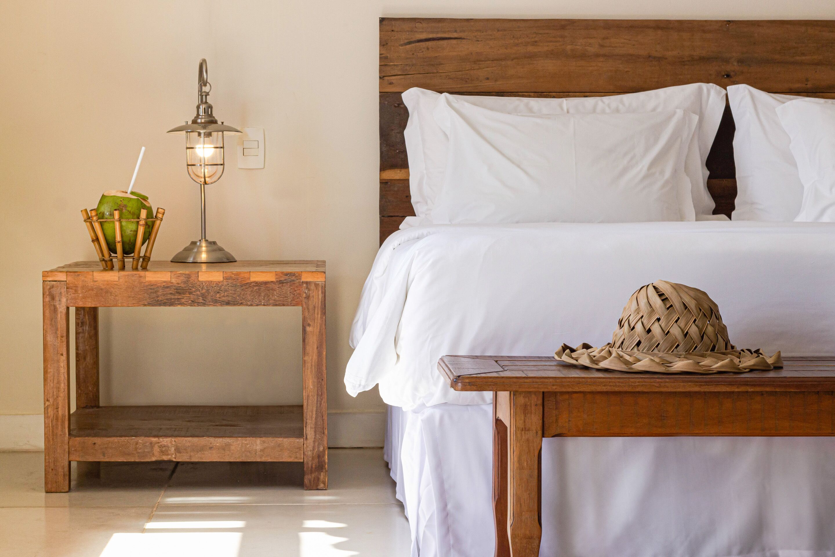 A neatly made bed with white linens, a wooden nightstand with a lamp and a coconut drink, and a woven hat on a table.
