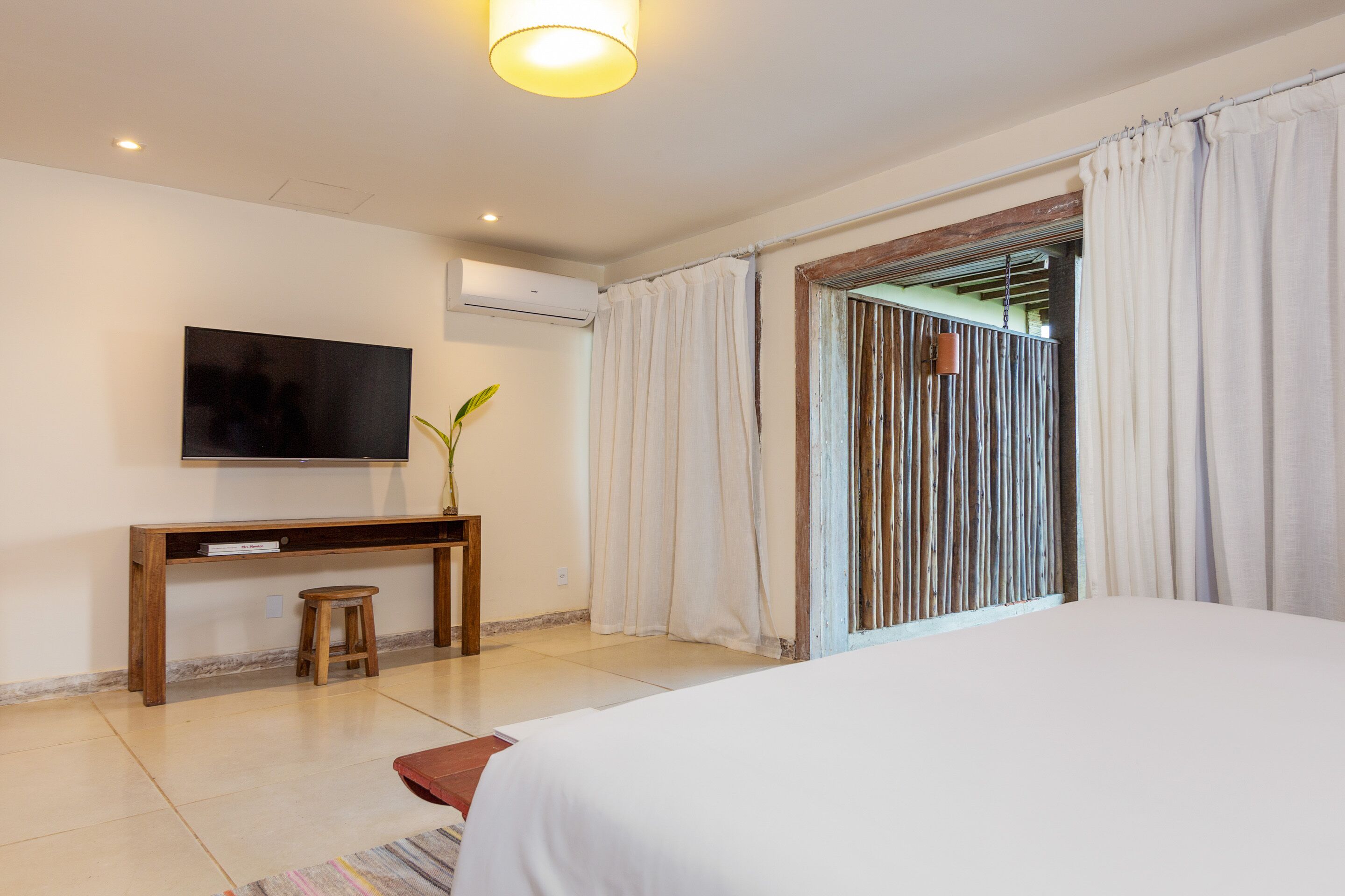 A bedroom with a bed, a TV mounted on the wall, a wooden console table, a small stool, and a window with curtains.