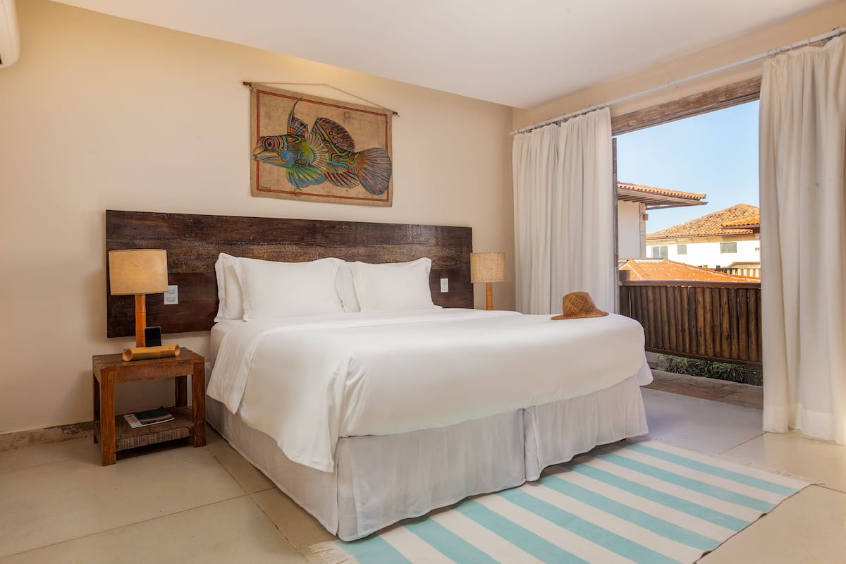A bedroom with a large bed, two bedside tables, a headboard, a wall-mounted artwork, and a view of the outside.