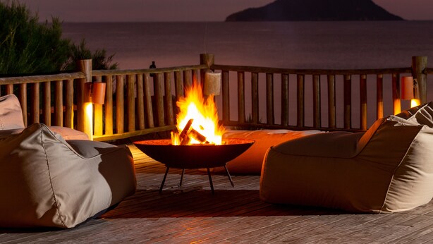 A deck with a fire pit, seating, and a view of the ocean at dusk. - Villa Rasa (Búzios)