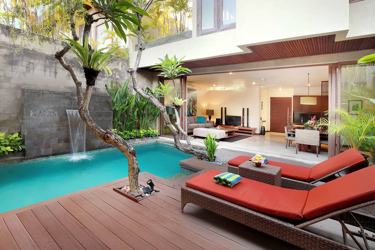 A modern outdoor pool area with a wooden deck, lounge chairs, and a waterfall feature.