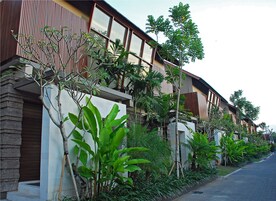 Um edifício residencial moderno com fachada combinando tijolo e madeira, grandes janelas e abundante vegetação, incluindo diversas plantas tropicais.
