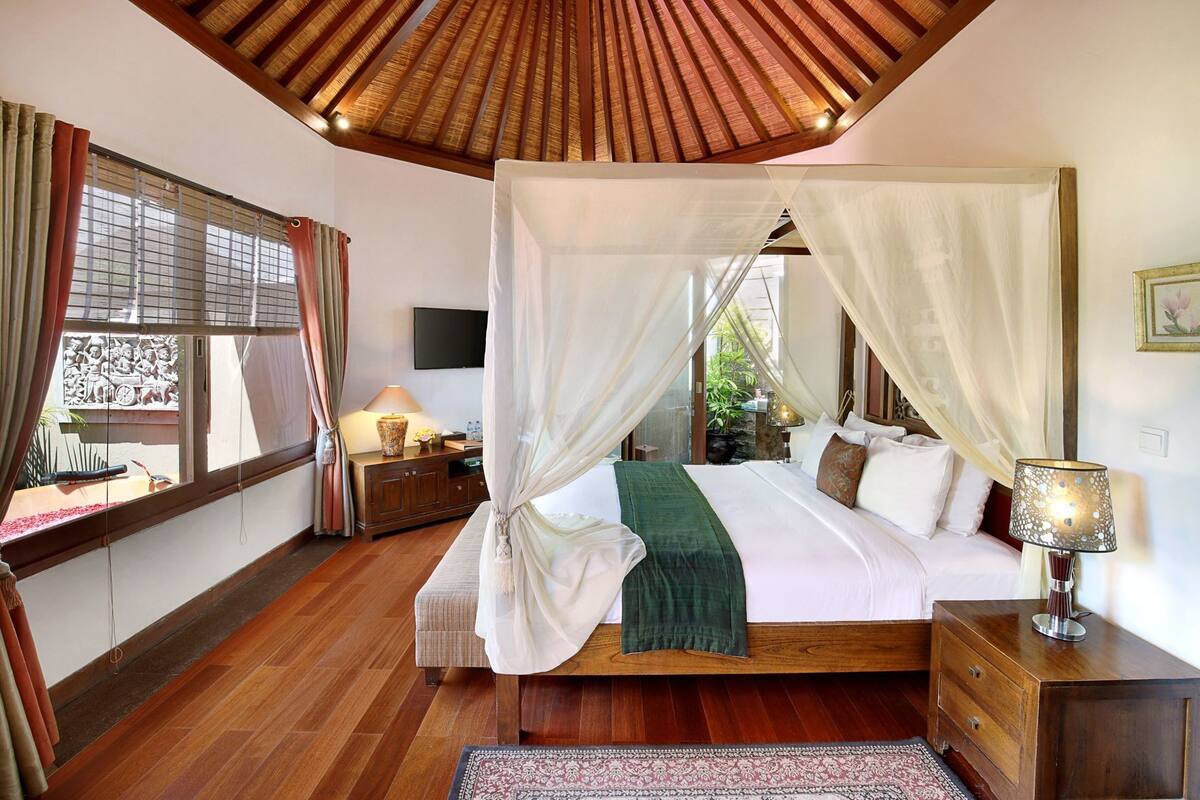 A bedroom with a wooden ceiling, a four-poster bed, a nightstand, a lamp, and a view of the outside.