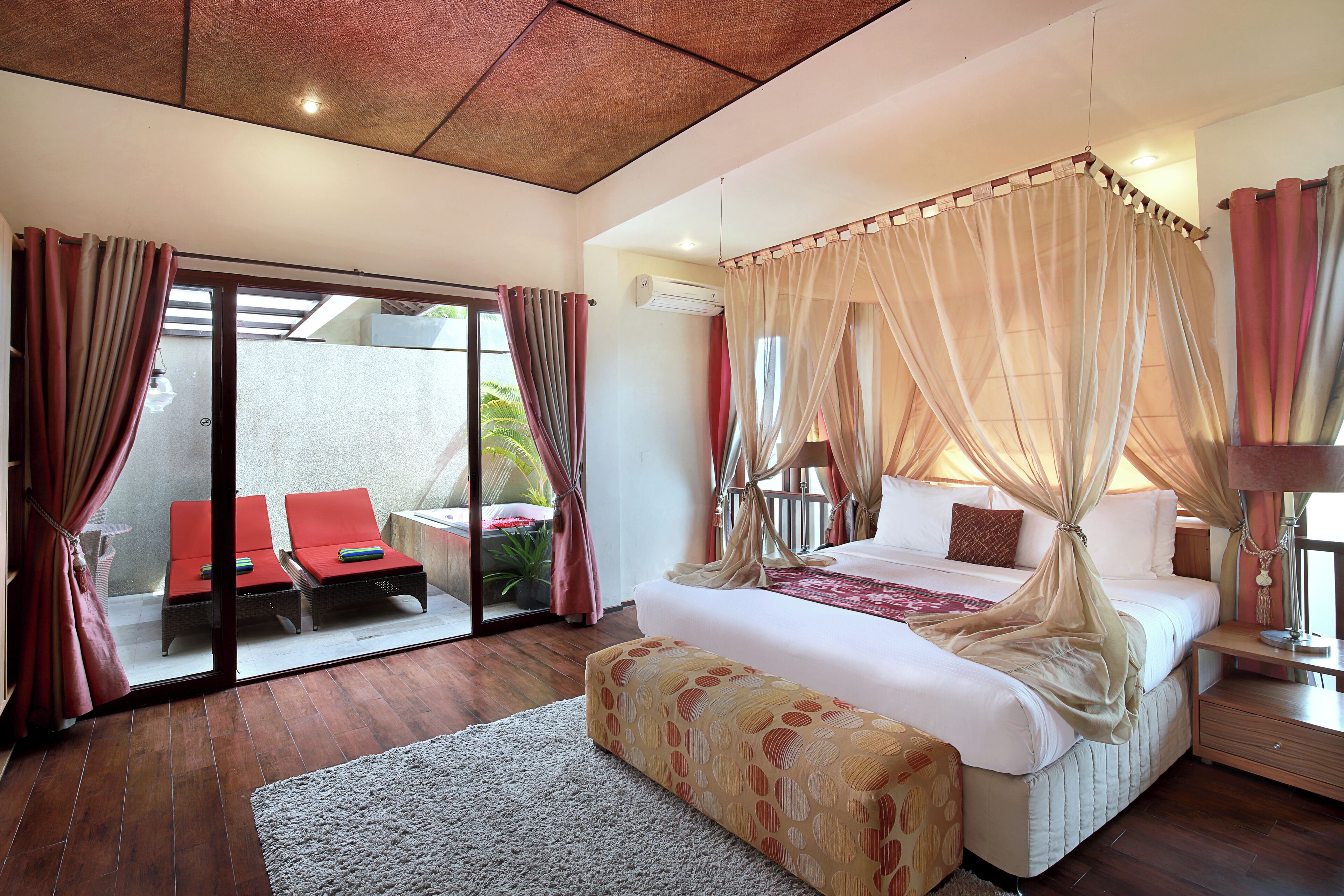 Une chambre à coucher avec un lit à baldaquin, un coin salon avec des fauteuils rouges et une vue sur l’extérieur, visible à travers une porte vitrée.
