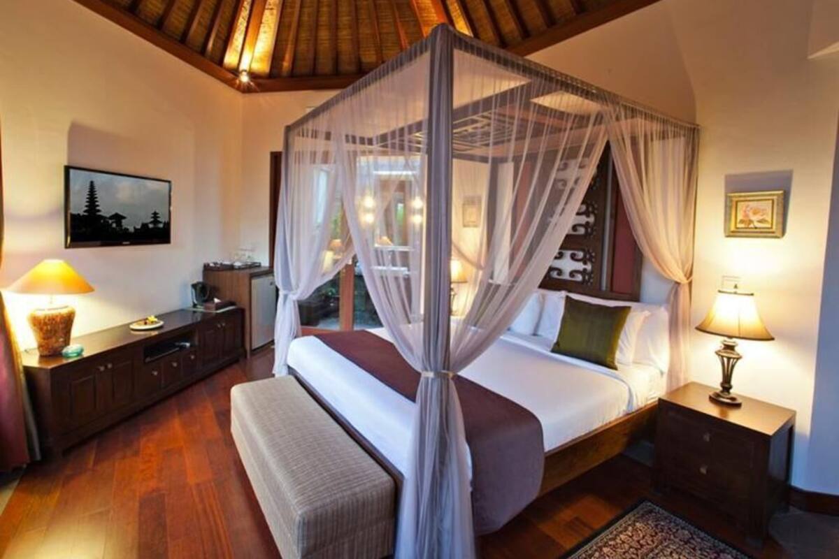 A bedroom with a four-poster canopy bed, wooden flooring, a nightstand with a lamp, a bench, and a framed picture on the wall.