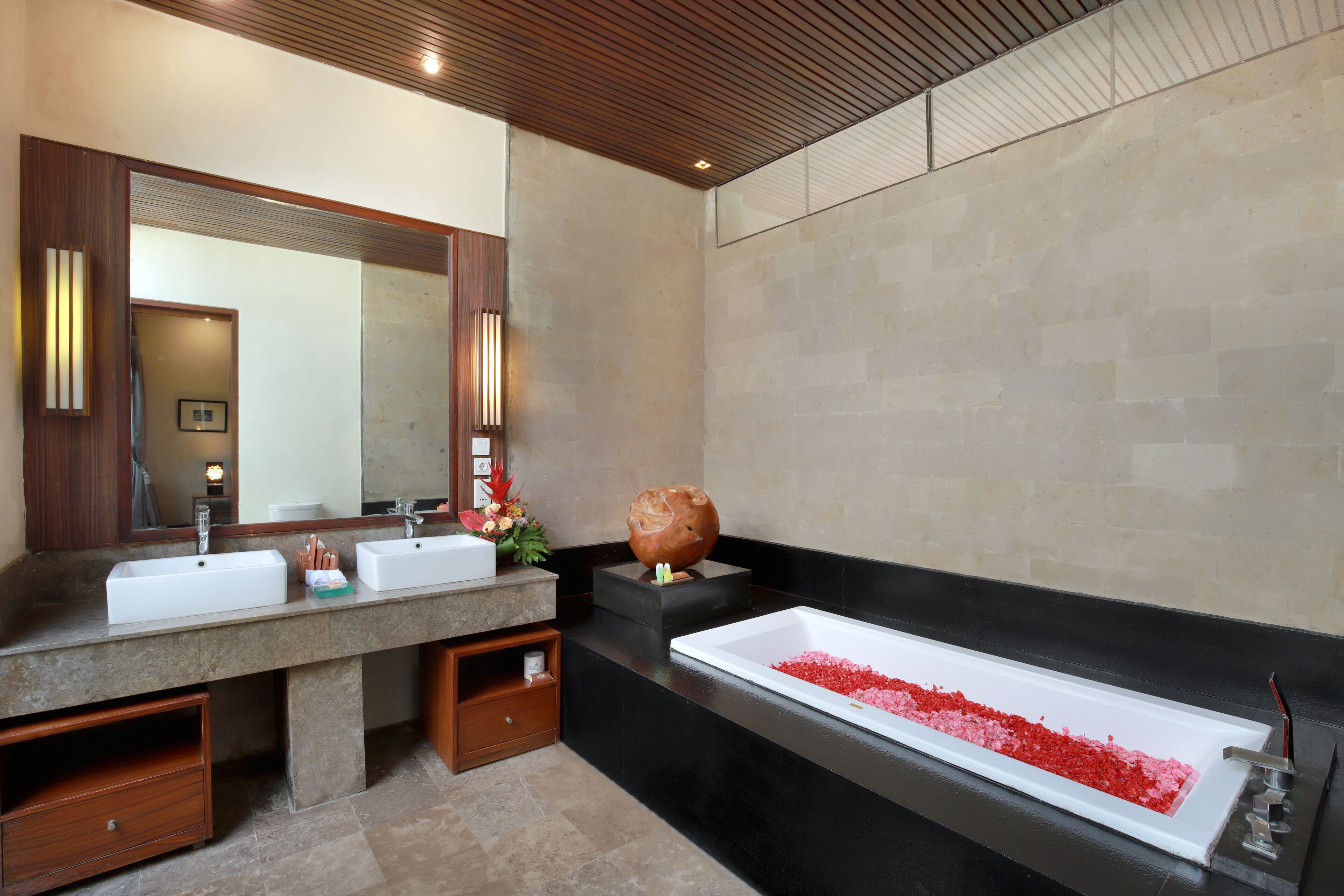Une salle de bain dotée d’un meuble-lavabo à deux vasques, d’une grande baignoire avec des sels de bain rouges et d’un plafond en bois.