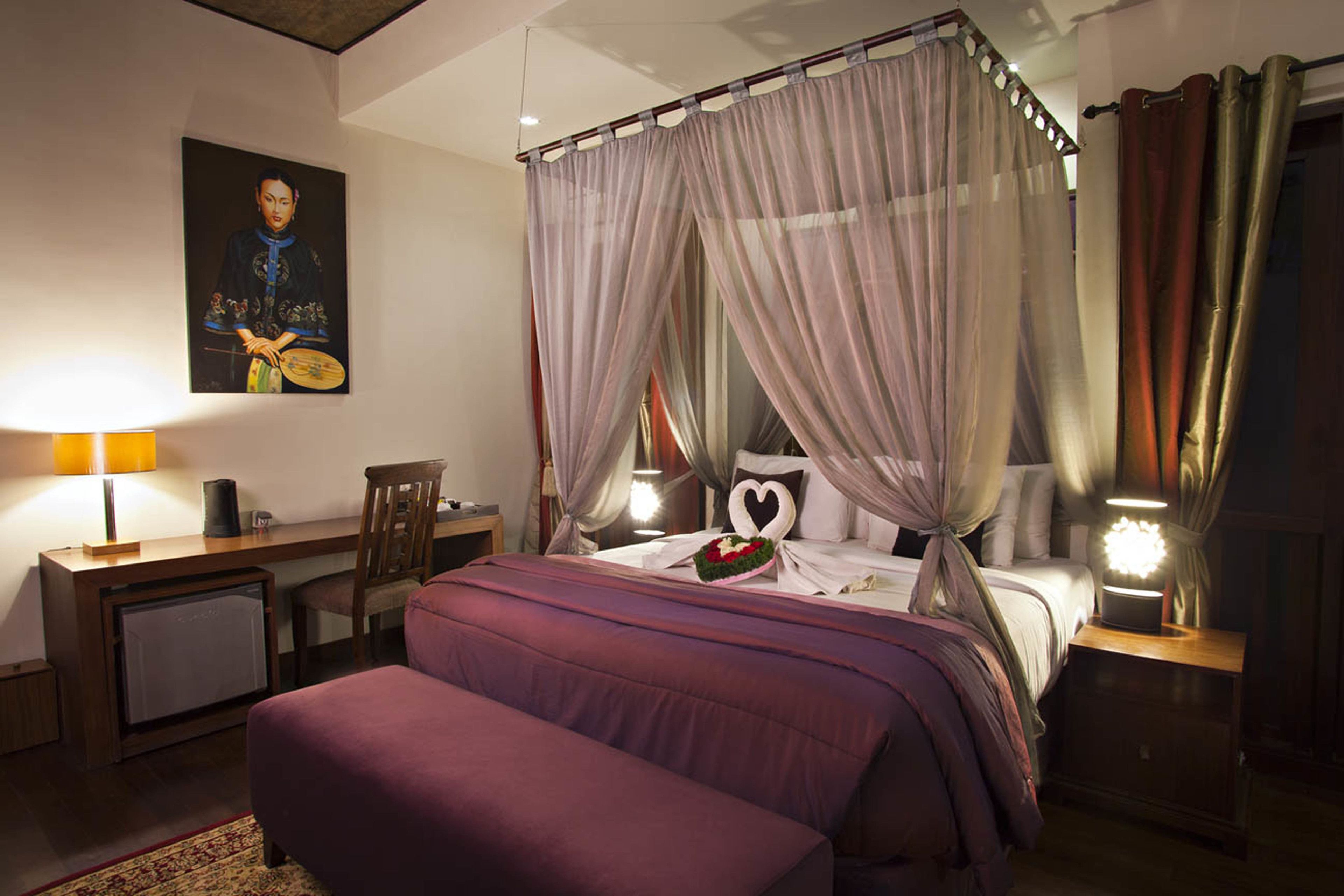 A bedroom with a four-poster bed, a desk with a chair, a bedside table with a lamp, and a wall-mounted painting.