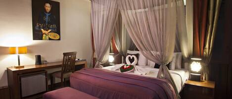 A bedroom with a four-poster bed, a desk with a chair, a bedside table with a lamp, and a wall-mounted painting.