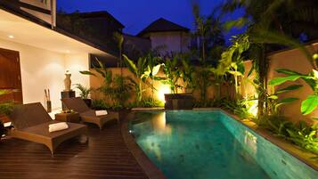A swimming pool with lounge chairs and a wooden deck, surrounded by tropical plants and illuminated by ambient lighting.