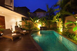 A swimming pool with lounge chairs and a wooden deck, surrounded by tropical plants and illuminated by ambient lighting.