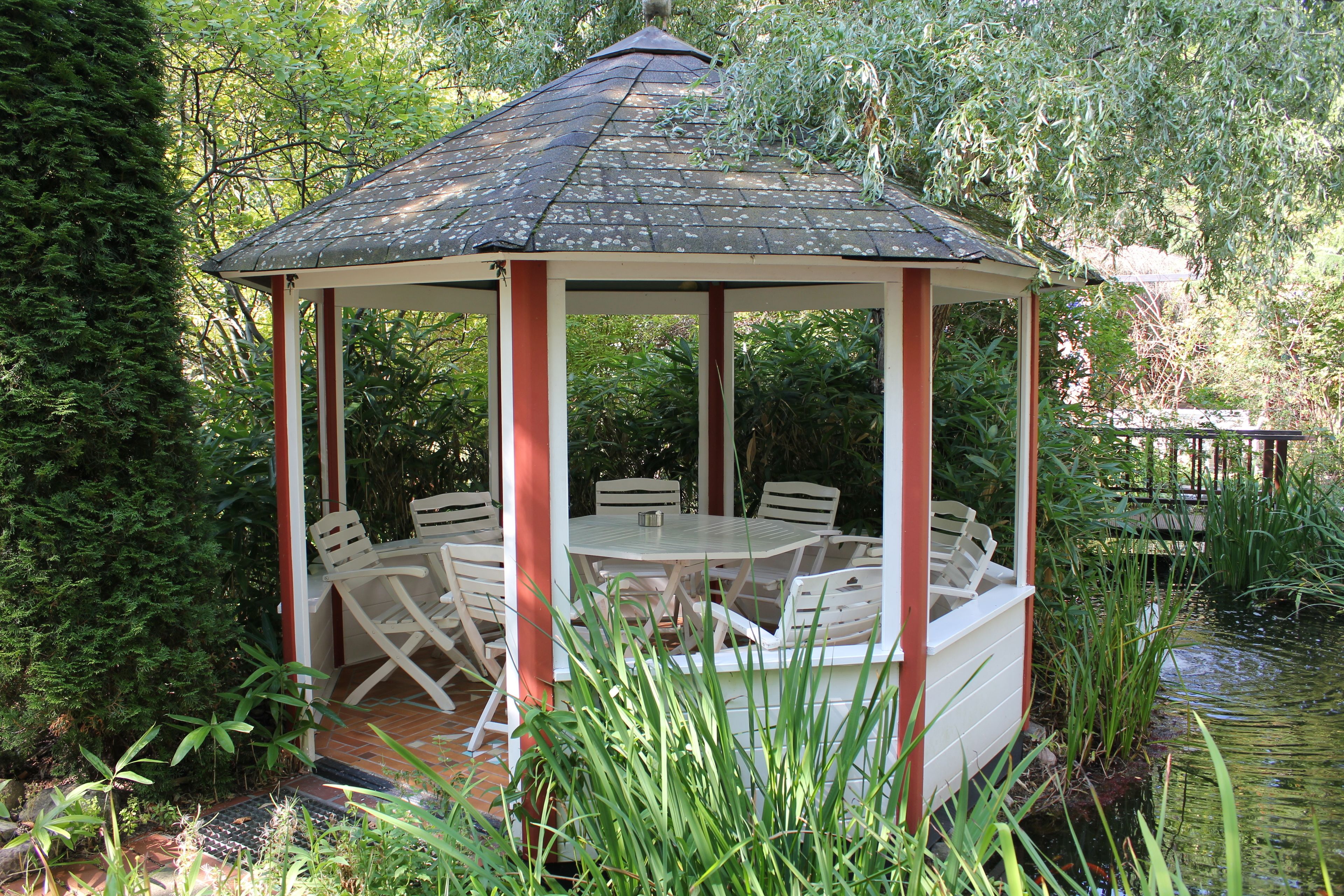 A gazebo with a tiled roof, surrounded by white chairs and a table, set in a lush garden with a pond.