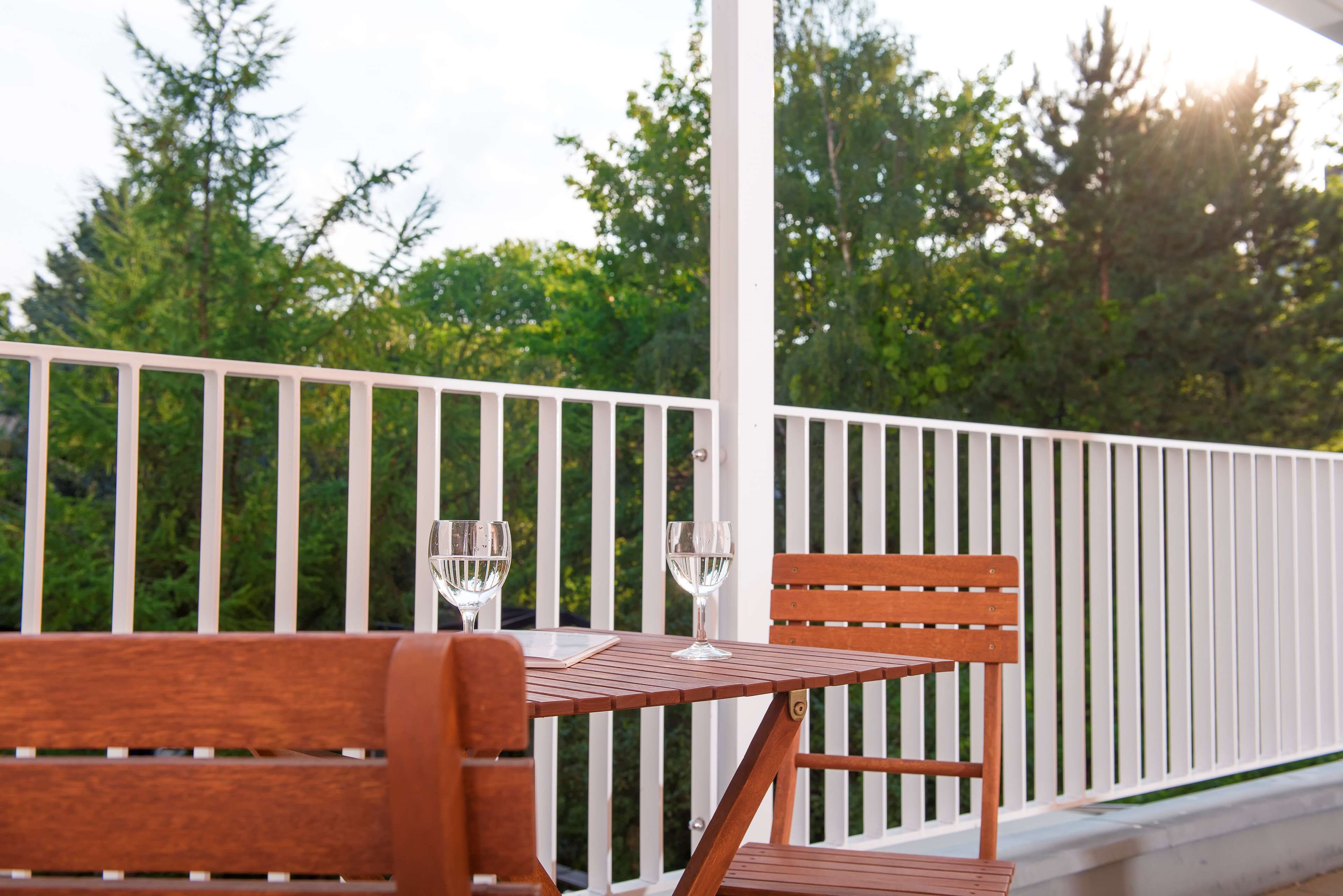 A wooden bench and table on a balcony with white railings, two glasses of water, and a view of trees and sky.