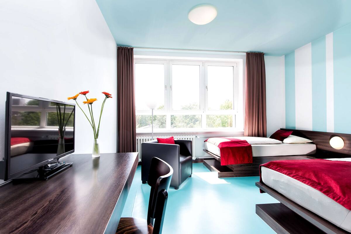 Habitación de hotel moderna con un ventanal grande, un escritorio con televisor de pantalla plana, un sofá y una cama con ropa de cama roja.
